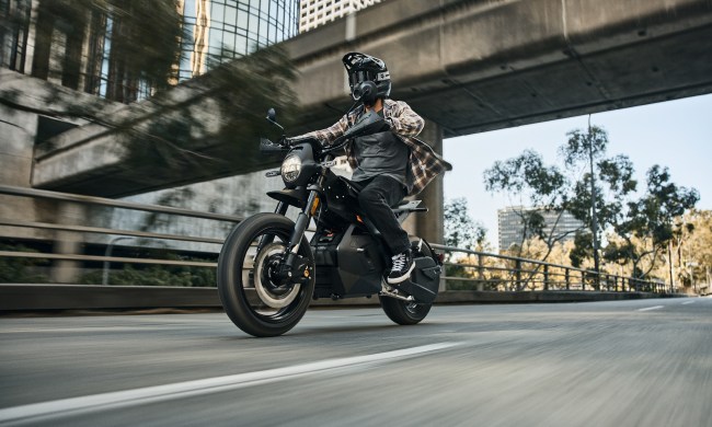 A rider on a Ryvid Outset electric motorcycle in Vapor Grey on a city street under an overpass.